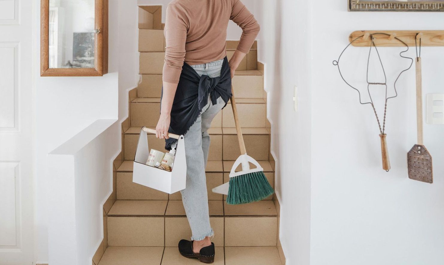 A woman carrying a cleaning caddy and broom up tiled stairs in a home